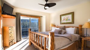 Une chambre avec un lit en bois, un ventilateur de plafond, une fenêtre avec des rideaux, une commode en bois et un tableau encadré accroché au mur.