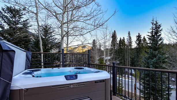 Un bain à remous sur une terrasse en bois, avec une rambarde en métal, entouré d’arbres et avec une montagne en arrière-plan.