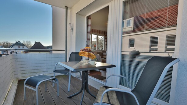 En balkong med ett bord och två stolar, ett fönster med persienner och utsikt över de omkringliggande husen.
