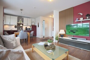A modern living room with a glass coffee table, a sofa, and a TV. There is a kitchen in the background with white cabinets and a dining area with a table and chairs.