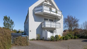 En modern byggnad i två våningar med en vit fasad, en balkong på övre våningen och ett litet inhägnat område med växter.
