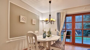 A dining area with a round table, white chairs, a chandelier, and a view of the outdoors through a large window. - Ferienparadies Klingberg Appartement 5 (Scharbeutz-Klingberg)