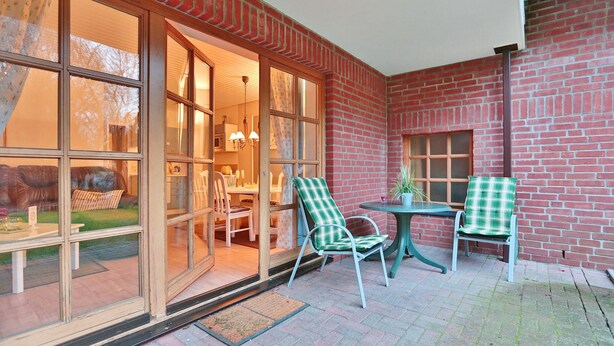 A patio area with two green chairs and a table, a brick wall, and a glass door leading to an interior space with a dining table and chairs. - Ferienparadies Klingberg Appartement 5 (Scharbeutz-Klingberg)