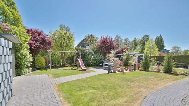 A backyard with a brick pathway, a swing set, a slide, and a pergola with potted plants.