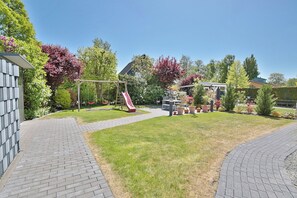 Un jardin avec un chemin en briques, une balançoire, un toboggan et une pergola ornée de plantes en pot.
