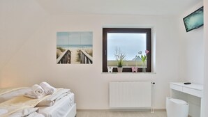 A bedroom with a bed, a window with a view of the beach, a radiator, and a TV on the wall.