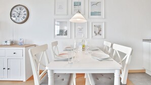 A dining area with a white table set for four, surrounded by white chairs, a hanging light fixture, and framed pictures on the wall. - Berliner Hof Apartment 215 (Scharbeutz)
