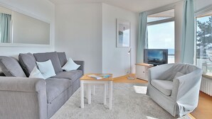 A modern living room with a grey sofa, a small wooden coffee table, a grey armchair, a television, and a large window with curtains. - Berliner Hof Apartment 215 (Scharbeutz)