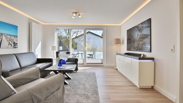 En moderne stue med sofa, sofabord og fjernsyn. Der er en skydedør, der fører ud til et udeområde med et bord og stole.