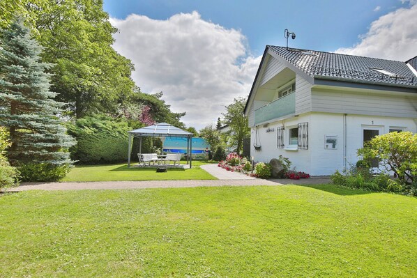 A two-story house with a white exterior and a tiled roof. There is a covered patio area with a blue umbrella and a bench. The house is surrounded by well-maintained lawns and various plants.