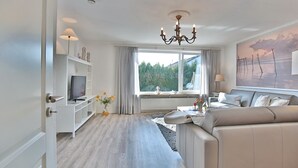 A modern living room with a sofa, TV, and a large window with curtains. - Appartementhaus Linda-lu Appartement 3 (Scharbeutz)