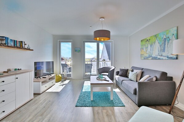A modern living room with a grey sofa, a TV, and a coffee table. There is a bookshelf with books and a painting on the wall. - Haus Meeresbrise Appartement 16 (Haffkrug)