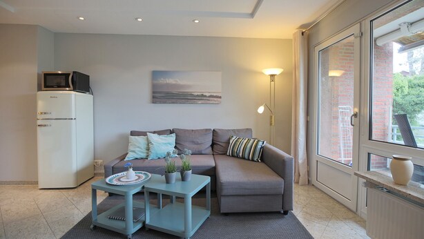 Een moderne woonkamer met een grijze bank, een klein tafeltje met decoratieve objecten, een witte koelkast en een groot raam met uitzicht.