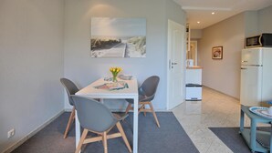 Een moderne keuken met een magnetron, een koelkast en een spoelbak. Er is een zithoek met een tafel en stoelen, en een schilderij van een zandstrand op de muur.