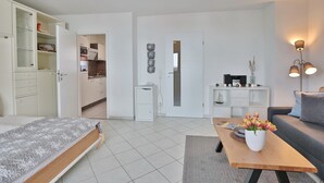 Een modern appartement met een bed, een slaapbank, een eettafel met een bloevas en een zichtbare keuken door een geopende deur.