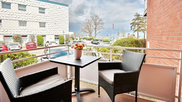 Een balkon met rieten meubilair en een tafel, met uitzicht op een parkeerplaats en gebouwen.