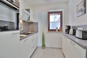 Une cuisine compacte avec des meubles blancs, un four à micro-ondes encastré, une machine à café et une fenêtre avec des stores.