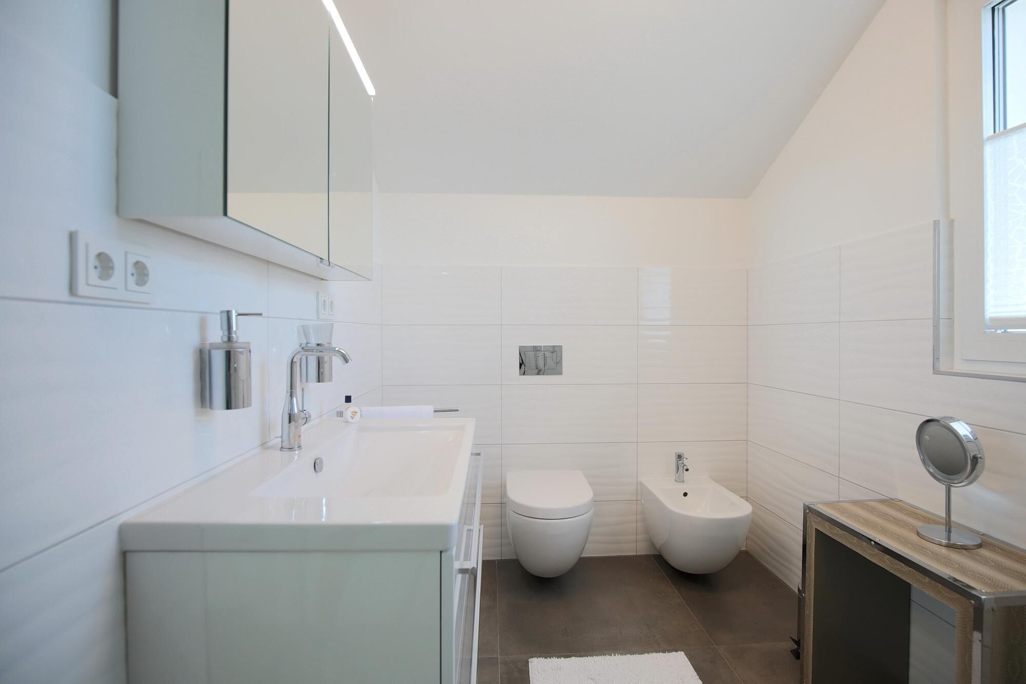 A modern bathroom with a white vanity, a toilet, a bidet, and a mirror.