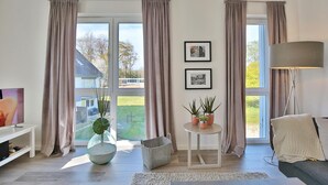 Un salon doté de grandes fenêtres, d’un téléviseur à écran plat, d’une table ronde agrémentée de plantes en pot et d’un lampadaire.