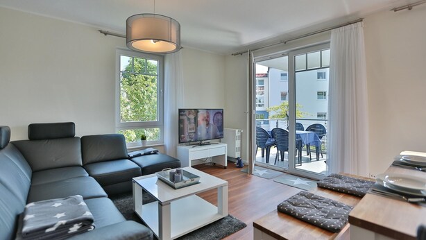 Een moderne woonkamer met een hoekbank, een flatscreen televisie, een salontafel en een eethoek met tafel en stoelen.