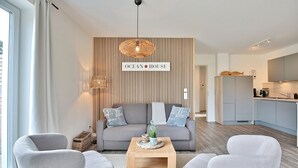 Moderni olohuone, jossa on sohva, kaksi nojatuolia, sohvapöytä ja seinälle kiinnitetty kyltti, jossa lukee: ”OCEAN HOUSE”.