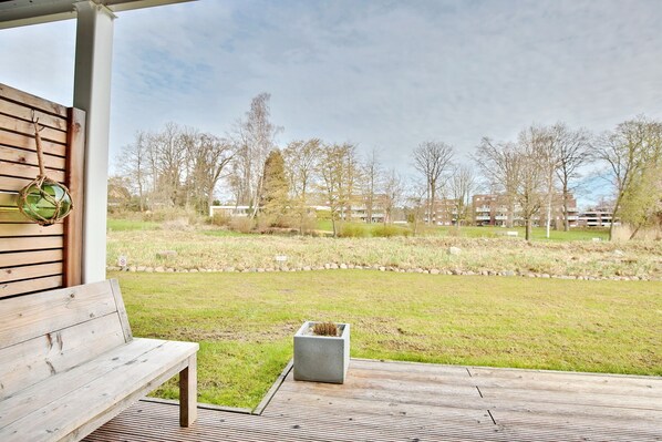 A wooden bench and a planter on a wooden deck with a view of a grassy area and buildings in the distance. - Haus Meeresbrise Appartement 1 (Haffkrug)