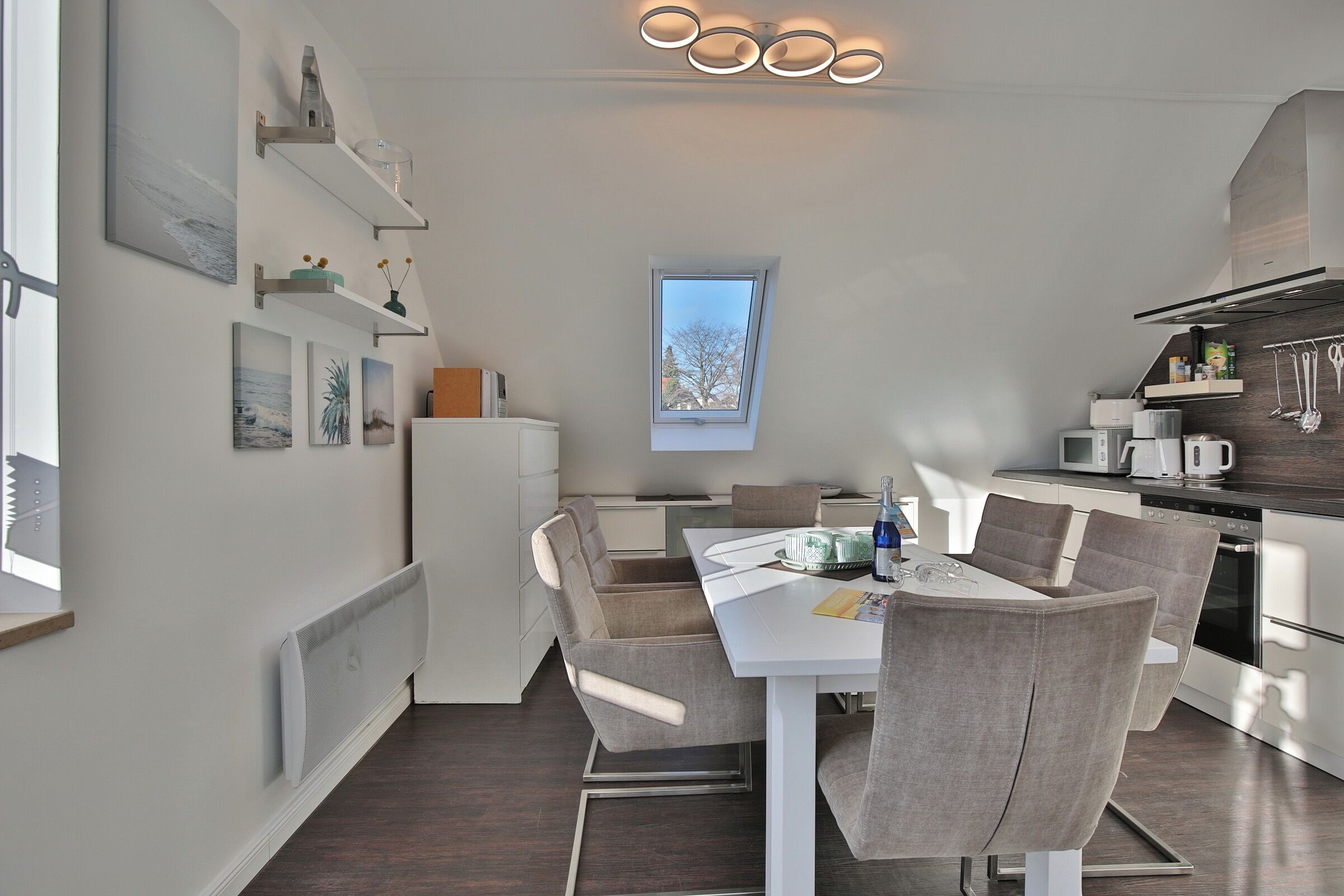A modern kitchen with a dining area, featuring a white table, grey chairs, and a window with a view.