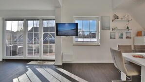 A spacious room with a large window, a flat-screen TV, and a dining area with chairs. - Parkdomizil Appartement 9 (Scharbeutz)