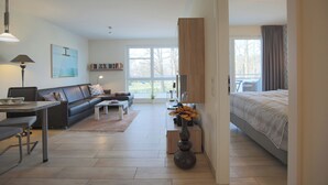 Sala de estar moderna com sofá, mesa de centro e área de jantar com mesa e cadeiras. Há uma janela grande que oferece luz natural e vista para o exterior.