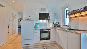 A modern kitchen with white cabinetry, a built-in oven, a microwave, and a large window providing natural light. - Ferienwohnung Dünenblick App. 1 (Scharbeutz)
