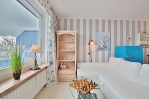 Uma sala de estar com um sofá branco, uma mesa de xadrez, um armário de madeira com copos, um refrigerador azul e uma janela com uma planta.