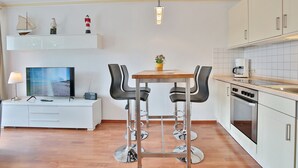A modern kitchen with white cabinets, a wooden table, two bar stools, a built-in oven, and a small shelf with decorative items. - Residenz Alte Bergstraße Appartement 14 (Scharbeutz)