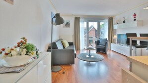 A modern living room with a sofa, a coffee table, and a TV. There is a window with curtains, a lamp, and a plant on the windowsill. - Residenz Alte Bergstraße Appartement 14 (Scharbeutz)