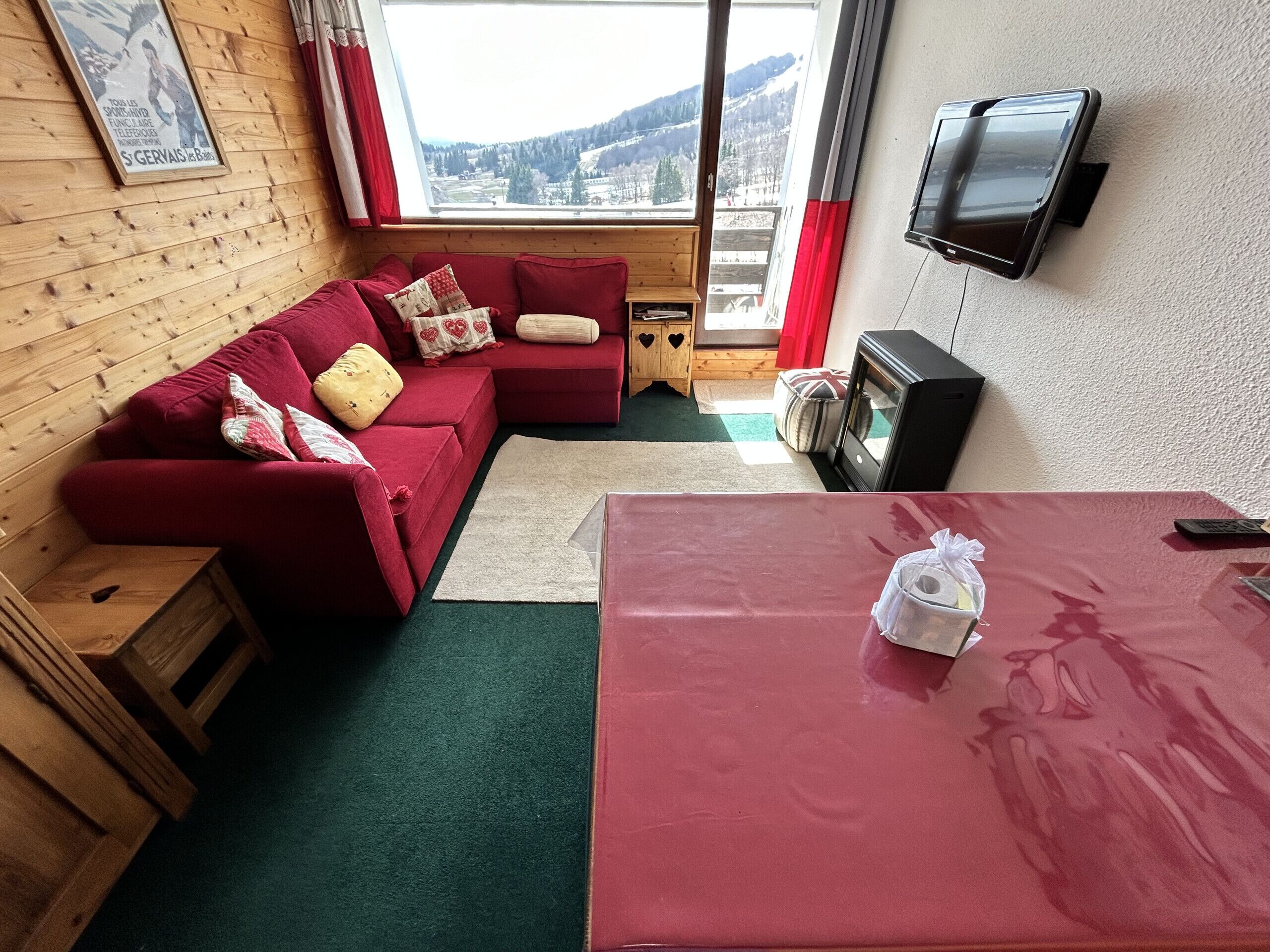 Una habitación acogedora con un sofá rojo, una mesita de madera y un televisor en la pared.
