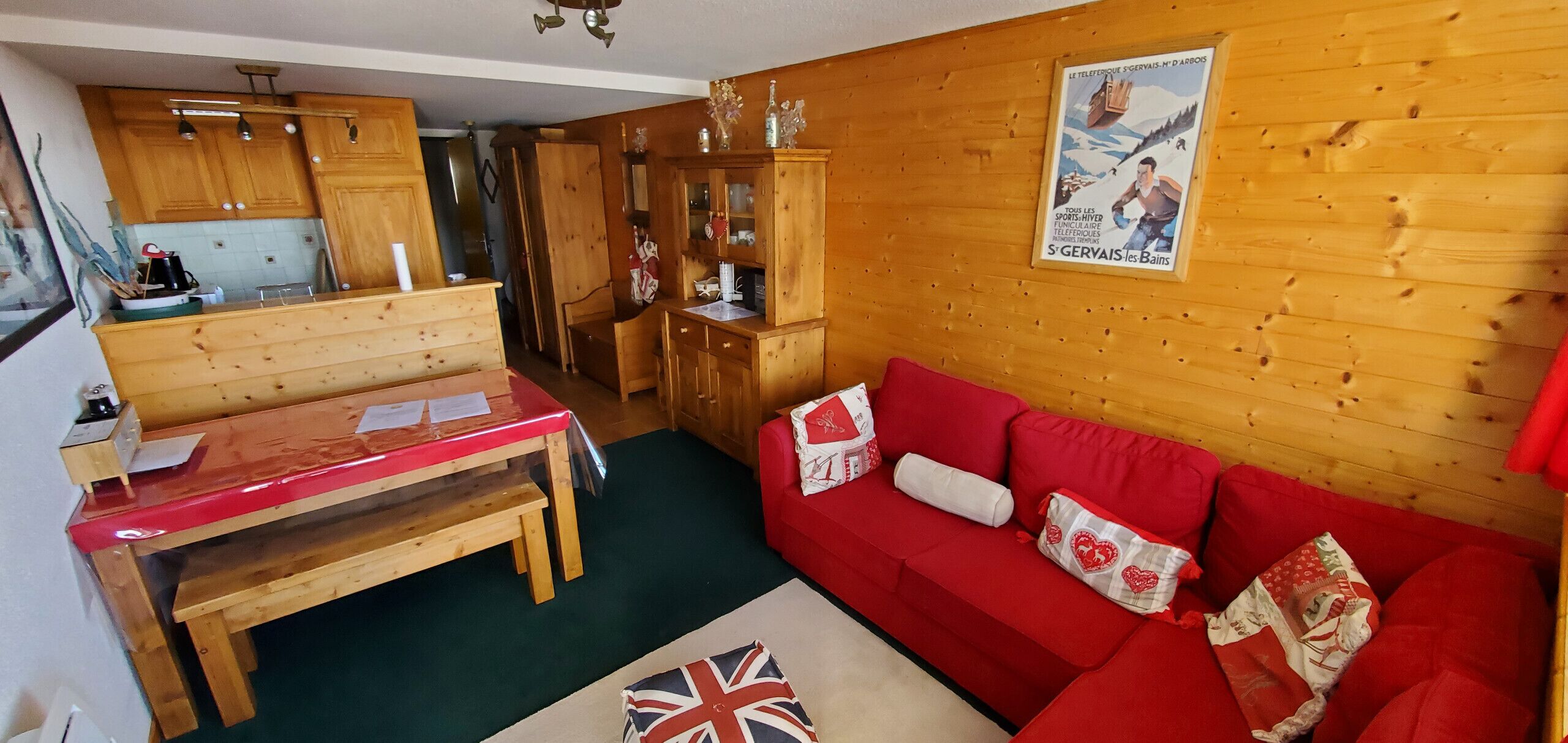 Un interno accogliente con un tavolo da pranzo in legno, un divano rosso con cuscini a motivi a cuore e un quadro appeso al muro.