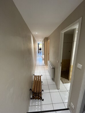 Een hal met een houten bankje, vloerbedekking van witte tegels en uitzicht op een badkamer met een douchecel.