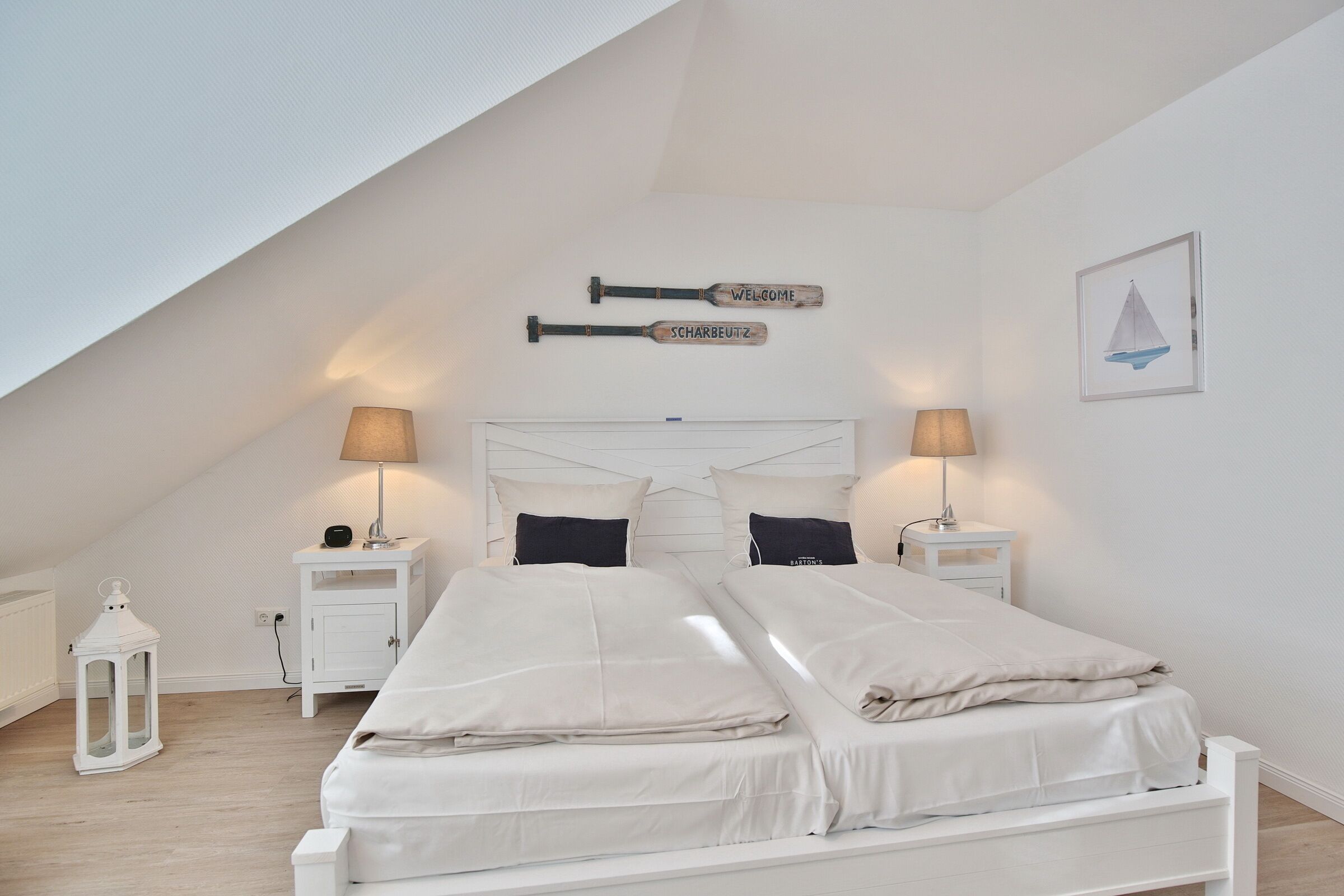 A bedroom with a sloped ceiling, a bed with white bedding, two bedside tables with lamps, a white lantern, and two wall-mounted signs.