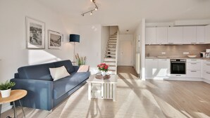 A modern living room with a blue sofa, a small coffee table with a vase of flowers, and a staircase leading to a kitchen with white cabinets. - Haus Seesand Hansastrasse 7, Penthouse Wohlfühlzeit (Scharbeutz)