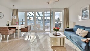A modern living room with a sofa, dining table, and chairs. There is a large window with a view of the outside. - Haus Seesand Hansastrasse 7, Penthouse Wohlfühlzeit (Scharbeutz)