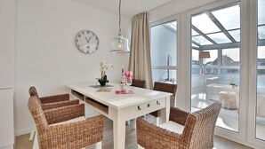 A dining area with a white table, wicker chairs, a clock, and a view through large windows. - Haus Seesand Hansastrasse 7, Penthouse Wohlfühlzeit (Scharbeutz)