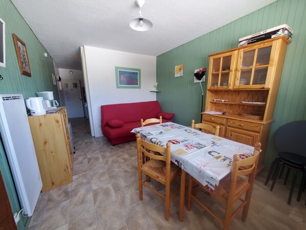 Een compacte kamer met een eettafel, stoelen, een rode bank, een houten kast en een kitchenette.