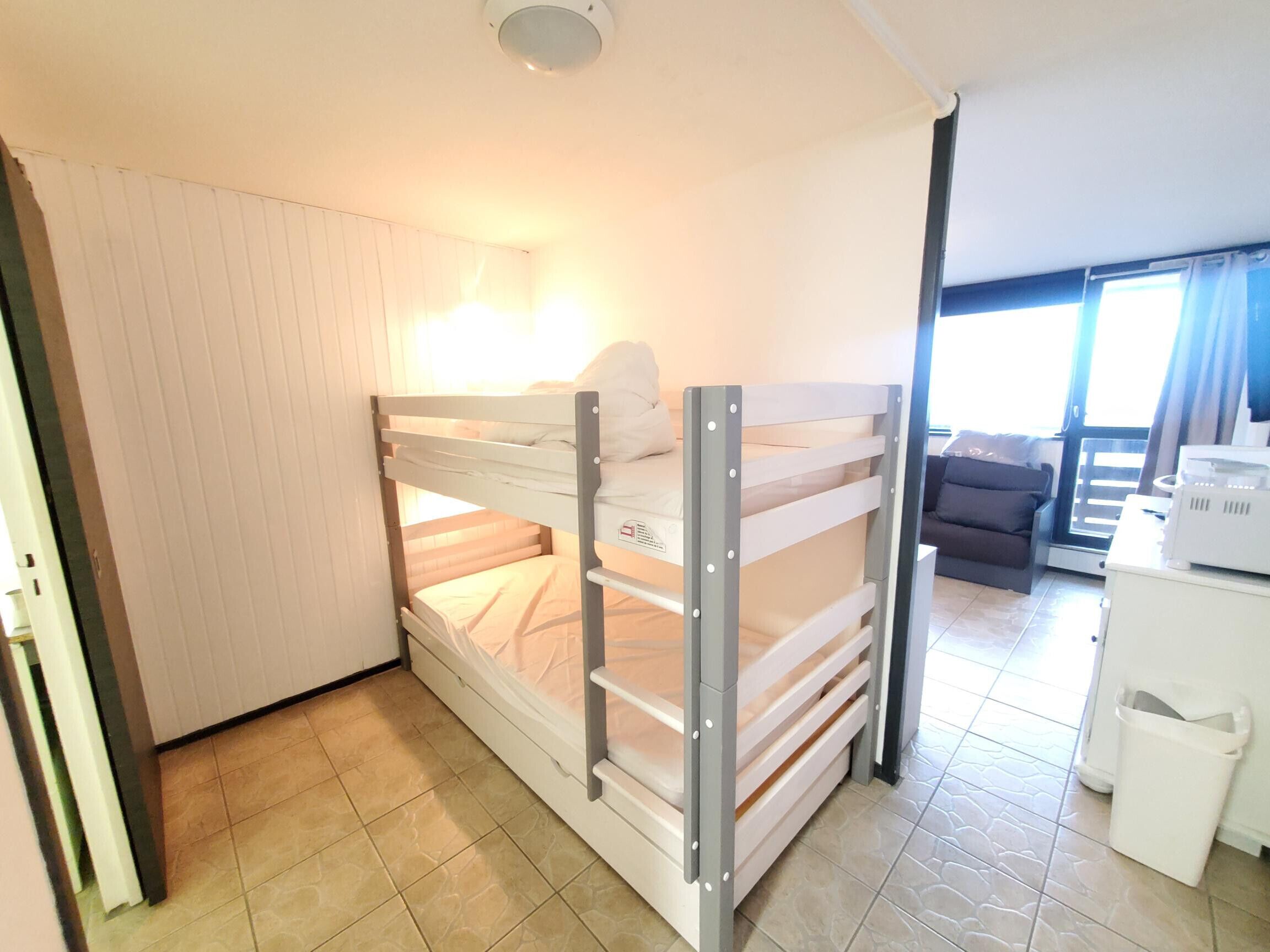 Una cama individual superpuesta con escritorio y silla, una ventana con cortinas, y suelo de baldosas.