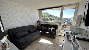 Ein modernes Wohnzimmer mit einem dunkelgrauen Sofa, einem kleinen Tisch mit Wasserkocher und Tasse sowie einem großen Fenster mit Blick auf eine gebirgige Landschaft.