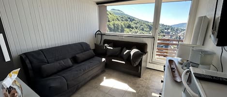 Una sala de estar moderna con un sofá gris oscuro, una mesita con una tetera y una taza, y un ventanal con vistas a un paisaje montañoso.