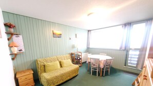 Um quarto pequeno e aconchegante com parede verde, sofá amarelo estampado, mesa lateral de madeira, cômoda de madeira pequena, mesa redonda com cadeiras e janela com cortinas.