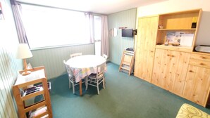 Um quarto com uma mesa redonda, cadeiras, um armário de madeira, uma televisão e uma janela com cortinas.