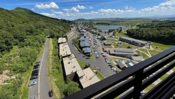 Um estacionamento com várias filas de carros, uma estrada que leva a um complexo de edifícios e colinas verdes ao fundo, sob um céu claro.