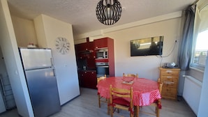 A compact kitchen with a refrigerator, microwave, and oven. There's a dining table with chairs and a clock on the wall.