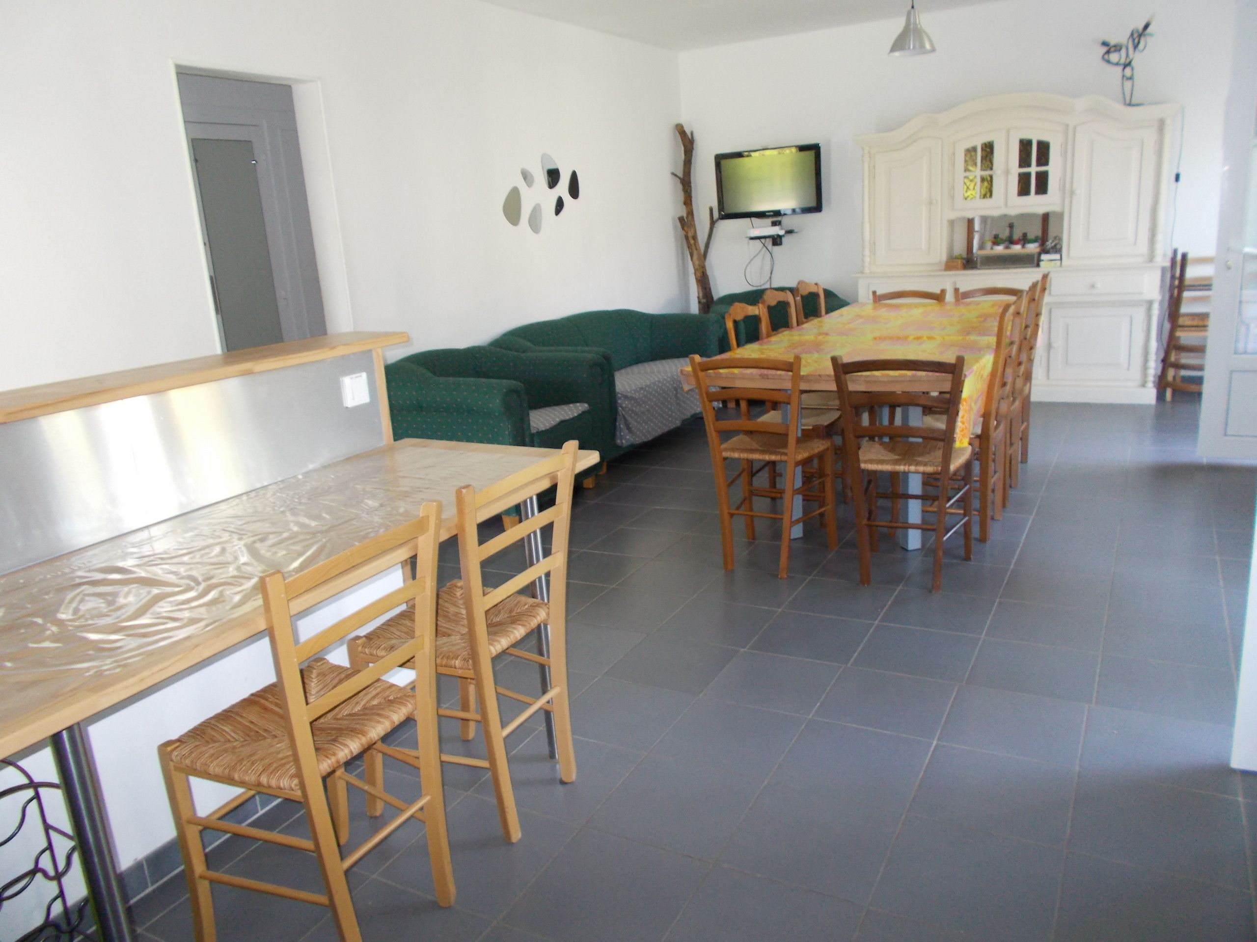 A dining area with a long wooden table and chairs, a green sofa, and a television mounted on the wall.