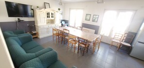 Uma sala de estar com sofá, uma mesa de jantar com cadeiras, TV fixada na parede e uma área de cozinha com armários e geladeira.